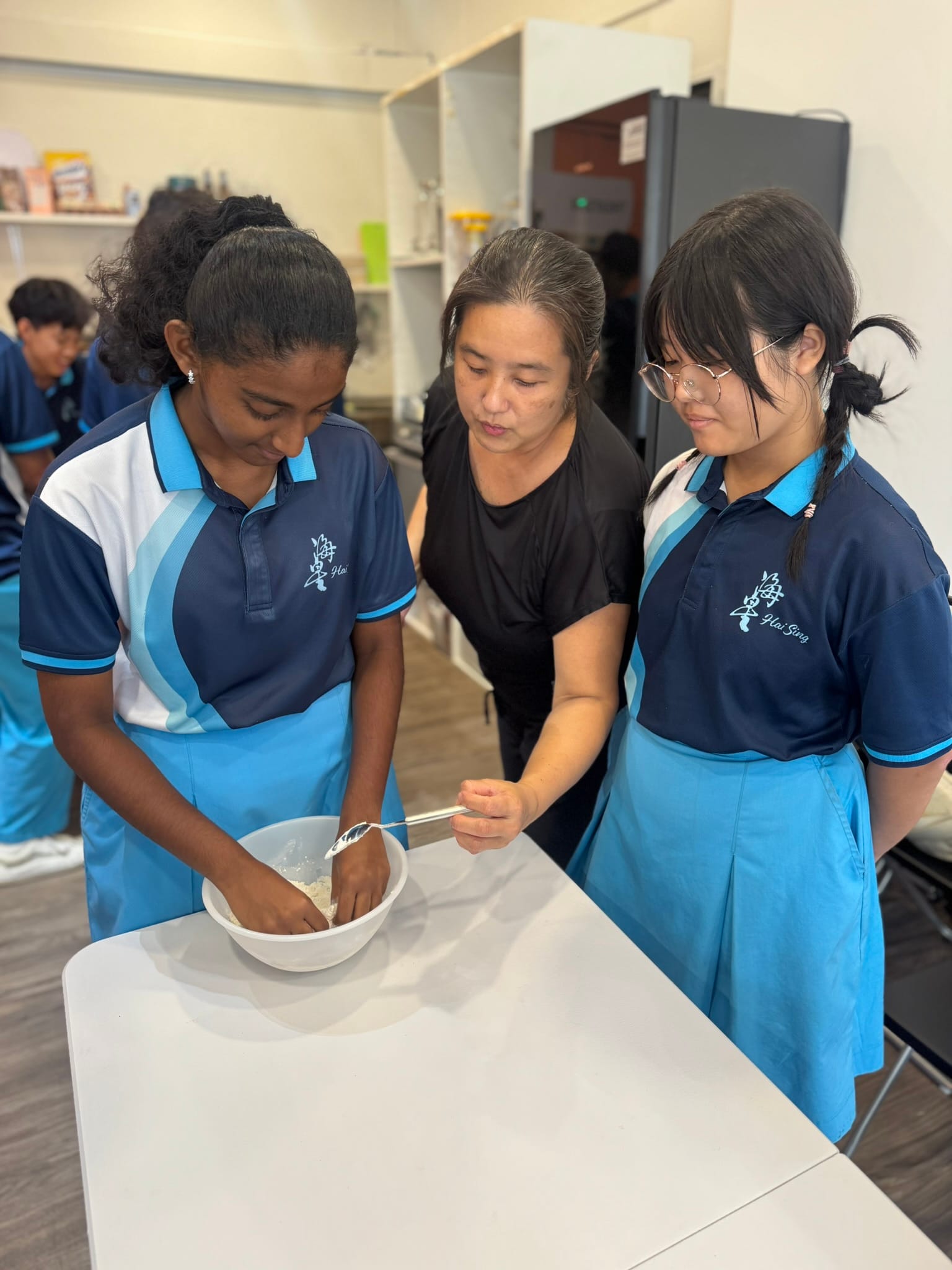 A teacher guiding two students in school uniform during a hands-on Nutrition & Food Science practical lesson.